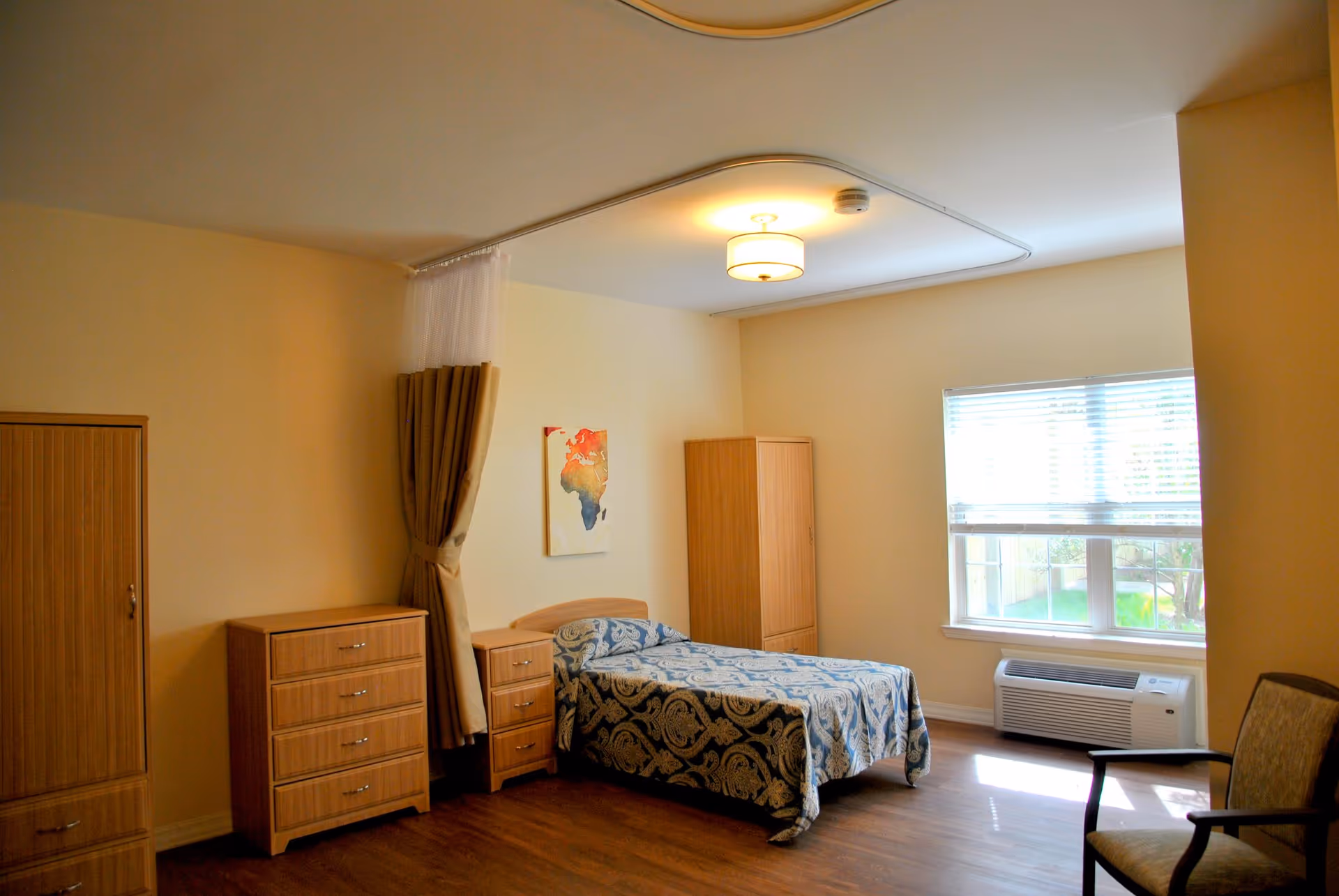 A simple and clean bedroom in an assisted living facility with a single bed covered in a blue patterned bedspread. The room has wooden furniture including a dresser, nightstand, and wardrobe. There is a window with blinds letting in natural light, a chair in the corner, and a ceiling light fixture. A curtain track is installed on the ceiling, partially holding a beige privacy curtain.