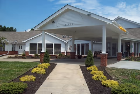 Front exterior view of a single-story senior living facility with a covered entrance supported by white columns with brick bases, landscaped with small shrubs and yellow plants along a concrete walkway leading to the entrance.