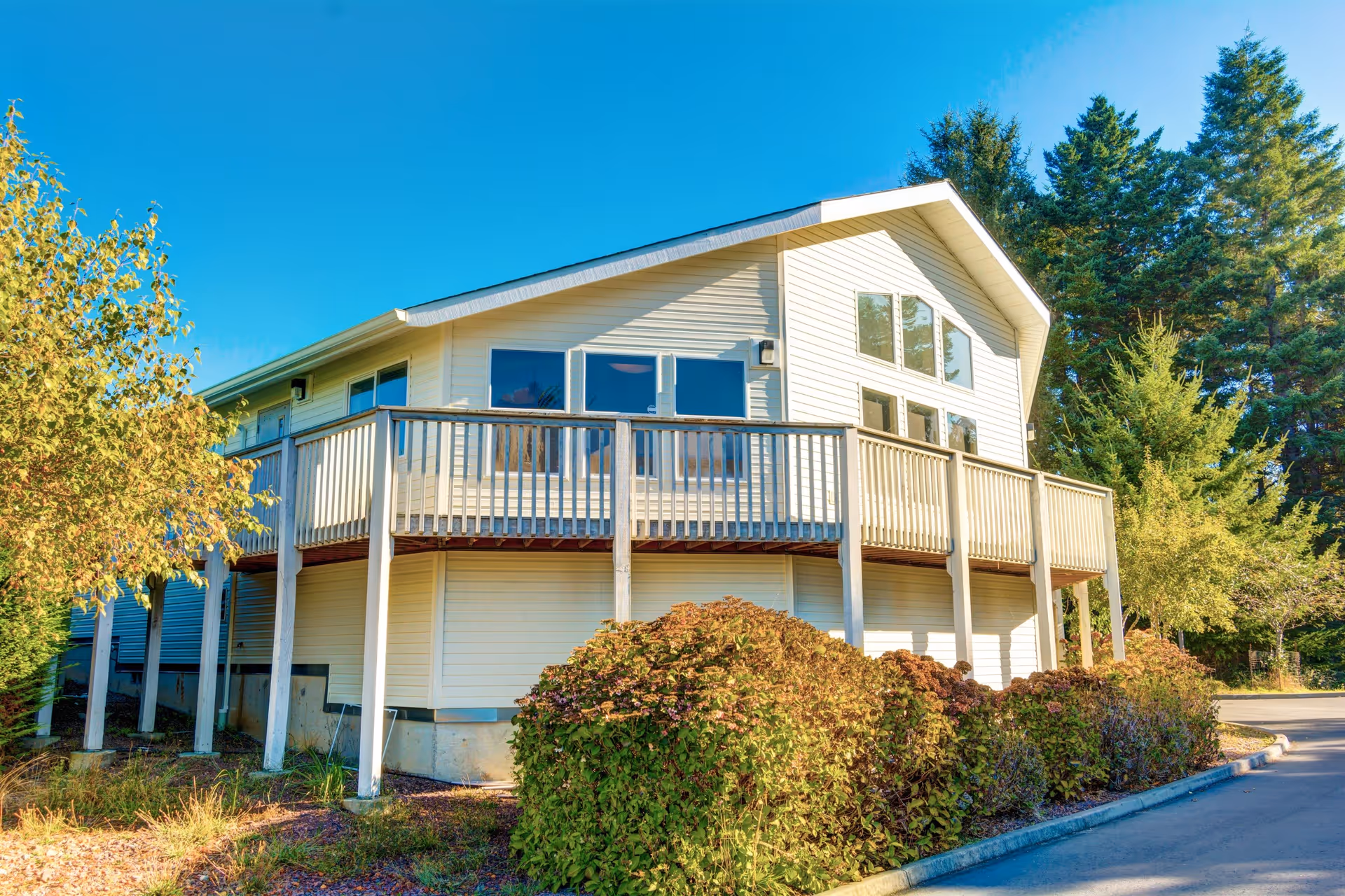 Exterior view of a two-story building with white siding and a wooden balcony railing, surrounded by bushes and trees under a clear blue sky.