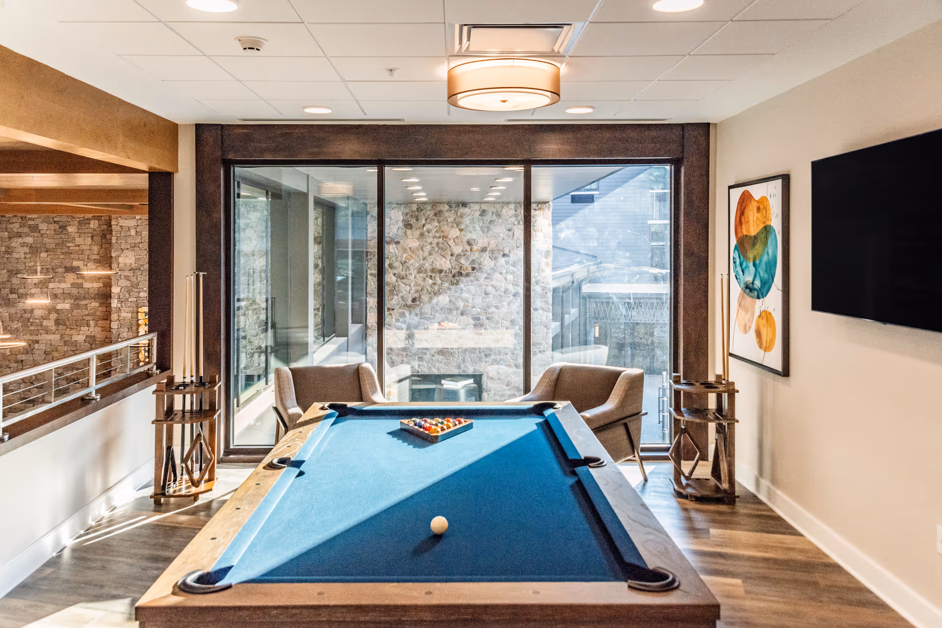 A bright recreational room featuring a pool table with a blue felt surface, two armchairs positioned near large windows with a stone wall view, a modern ceiling light fixture, a colorful abstract painting on the wall, and a large flat-screen TV mounted on the right wall.
