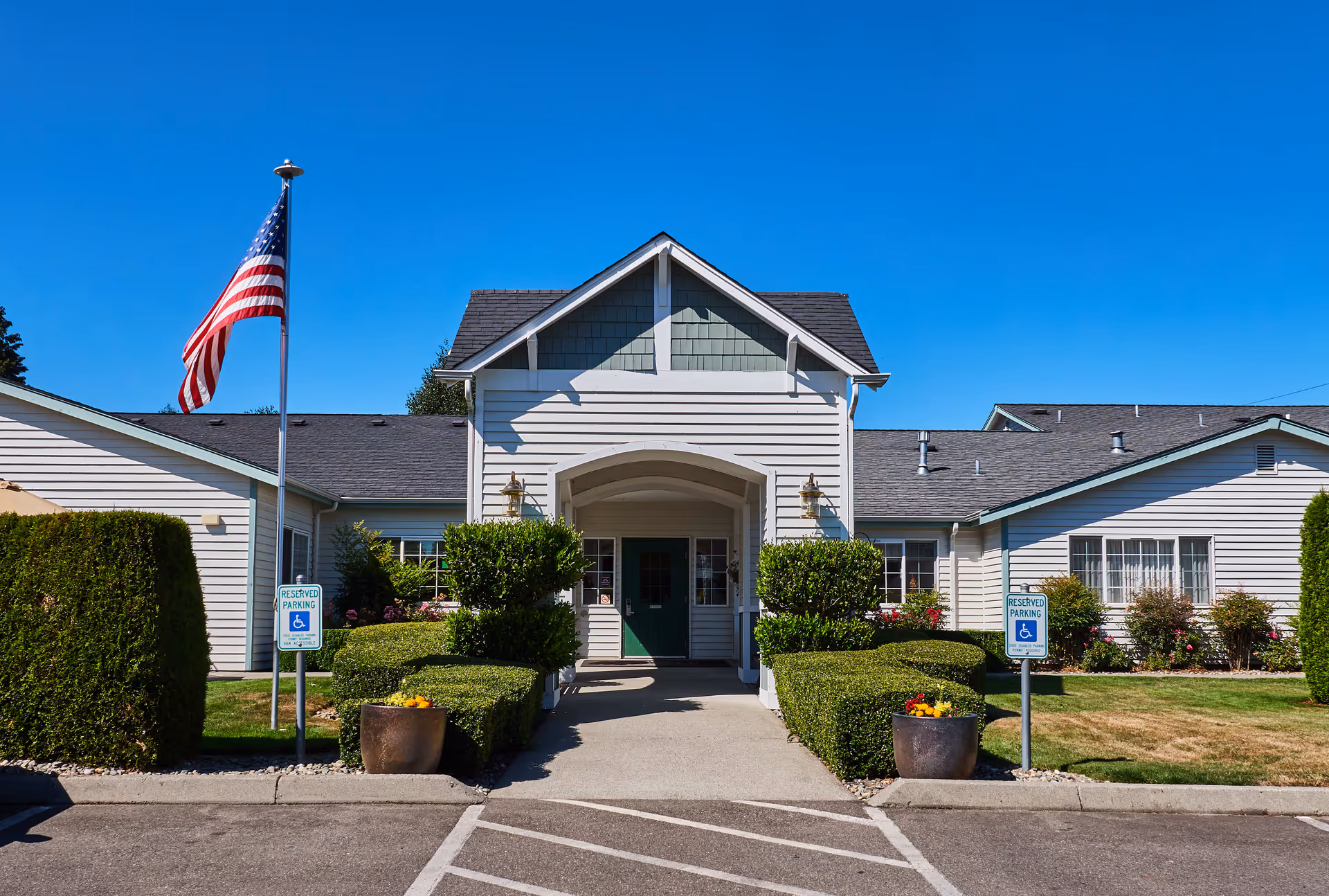 Front exterior view of a single-story assisted living facility building with white siding, a peaked roof, and a covered entrance. There are neatly trimmed bushes and flower pots on either side of the walkway leading to the entrance. An American flag is flying on a flagpole to the left, and two reserved parking signs for handicapped parking are visible near the entrance.