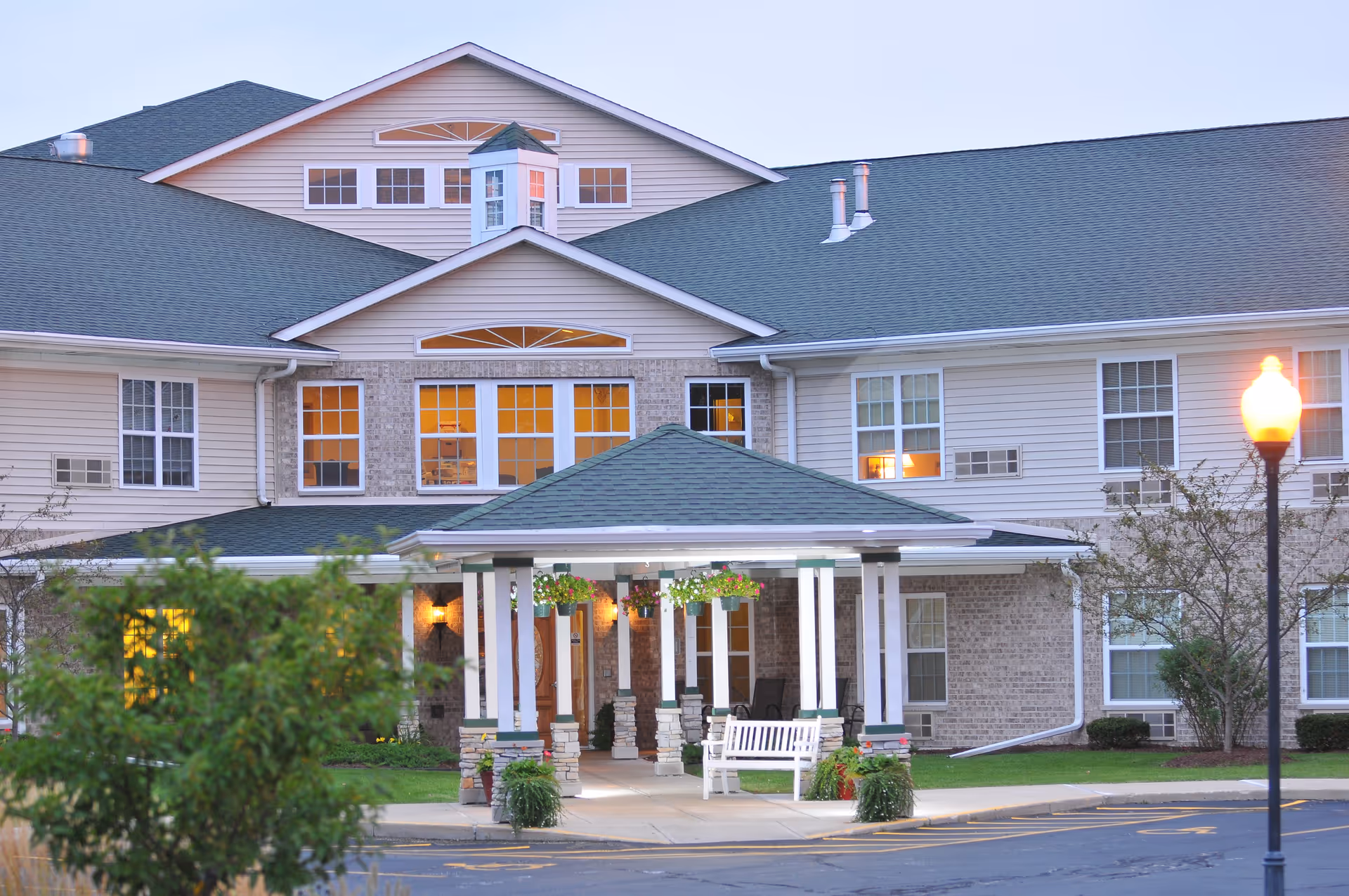 Front entrance of a multi-story assisted living building with a covered porte-cochere, benches, hanging plants and lit windows.