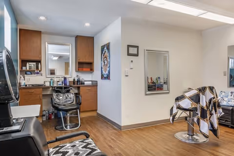 Interior view of a salon area in a senior living facility with two salon chairs, one covered with a patterned cape, a large mirror on the wall, wooden cabinets, and various hair care products on the counter.