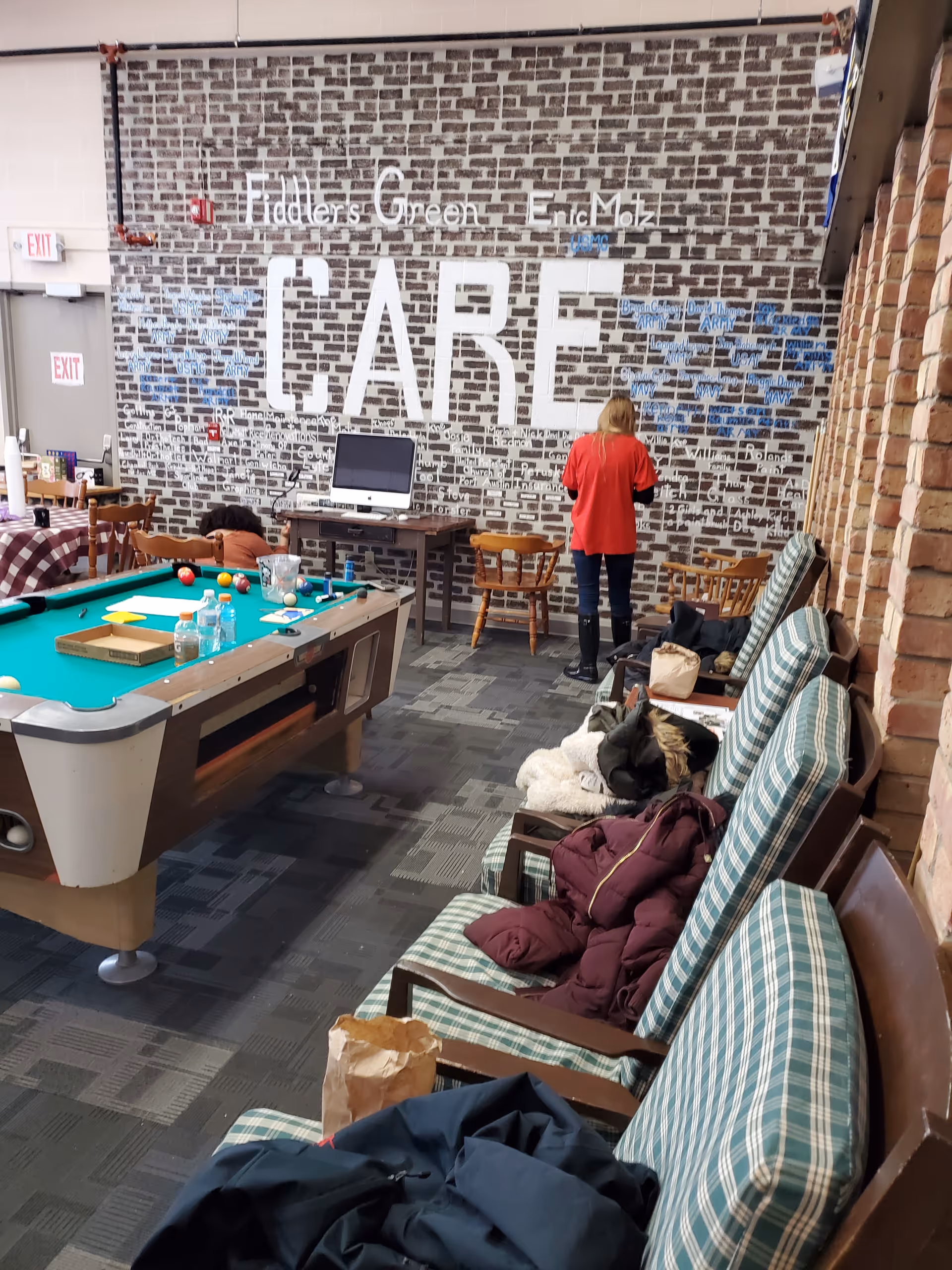 A communal recreation room with a pool table, cushioned chairs, a computer desk and a brick-patterned wall mural reading 'CARE'.
