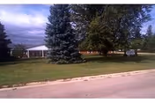 View of a single-story building partially obscured by large evergreen and deciduous trees, with a well-maintained grassy lawn in front and a paved road in the foreground. A sign is visible near the trees on the right side of the image.
