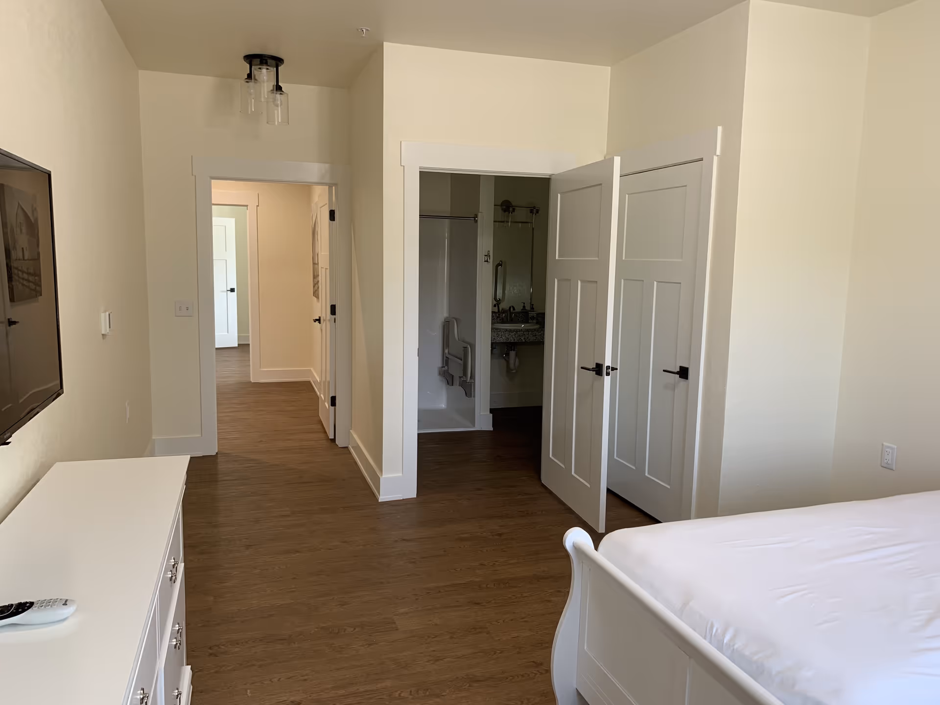 Bright furnished bedroom in an assisted living unit with a bed, dresser, wall-mounted TV, and an open doorway to a bathroom and hallway.