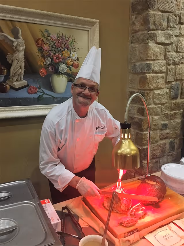 A chef wearing a white uniform and tall white chef hat is carving meat on a wooden cutting board under a heat lamp. Behind him is a stone wall and a framed painting of a vase with flowers and a statue. Plates and serving utensils are on the table nearby.