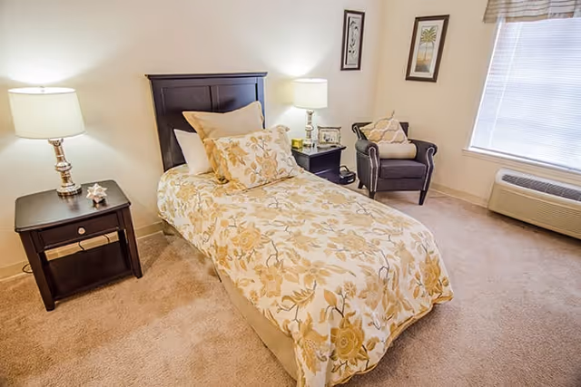 A cozy bedroom with a single bed covered in a floral patterned bedspread and multiple pillows. There are two dark wooden nightstands on either side of the bed, each with a lamp. A comfortable armchair with a decorative pillow is placed near a window with blinds. The room has beige carpeting and light-colored walls with framed artwork.