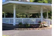 White gazebo with a lattice railing surrounded by greenery and flowers, situated on a paved area with trees in the background.