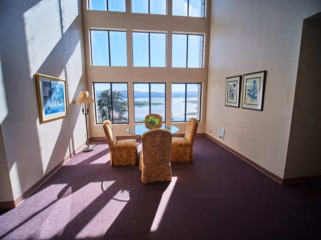 A bright room with large windows offering a scenic view of a river and landscape. Inside, there is a round glass table with four upholstered chairs arranged around it. The room has purple carpet, a floor lamp, and framed artwork on the walls.