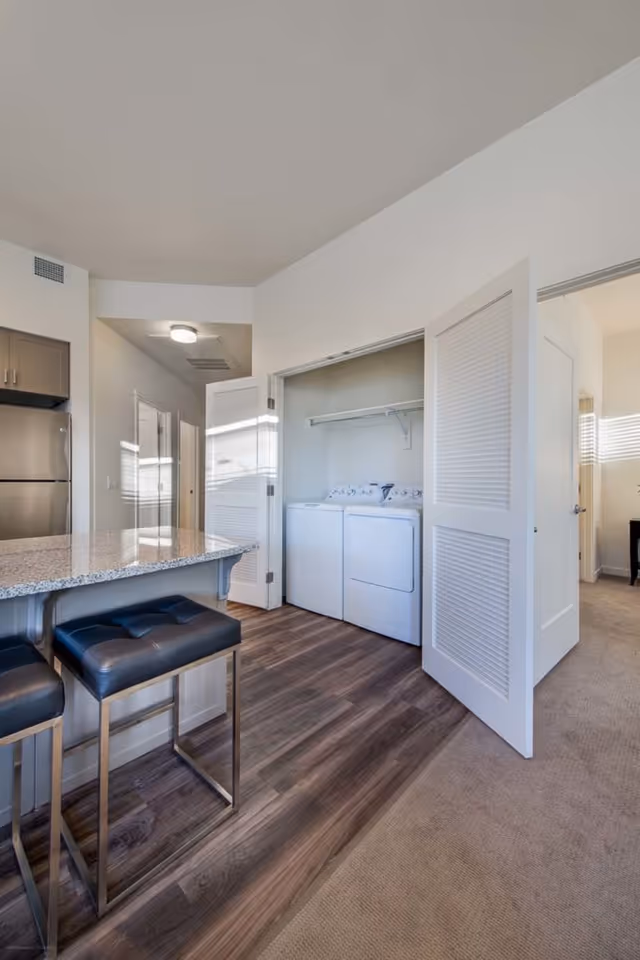 Interior view of a modern apartment showing a kitchen area with granite countertop and two black cushioned bar stools. In the background, there is a laundry closet with a washing machine and dryer behind open louvered doors. Adjacent rooms with carpeted floors are visible.