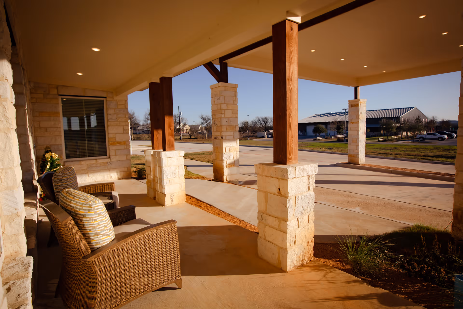 Covered outdoor patio area with stone pillars and wooden beams, featuring wicker chairs with cushions. The patio overlooks a driveway and a grassy area with a building and parked cars in the background under a clear blue sky.