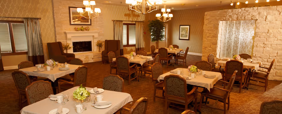 A cozy dining room with several tables covered with beige tablecloths, each set with plates, cups, and floral centerpieces. The room features patterned wallpaper, a stone fireplace with a framed picture above it, large windows with curtains, and warm lighting from chandeliers. Comfortable upholstered chairs surround the tables.