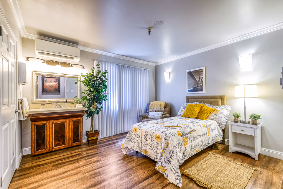 A cozy bedroom with a single bed covered in a floral bedspread with yellow pillows. Next to the bed is a white nightstand with a lamp and small plants. There is a comfortable armchair in the corner and a large window with vertical blinds. On the left side of the room, there is a wooden vanity with a sink, mirror, and a potted plant beside it. The room has wooden flooring and soft lighting from wall sconces and a floor lamp.