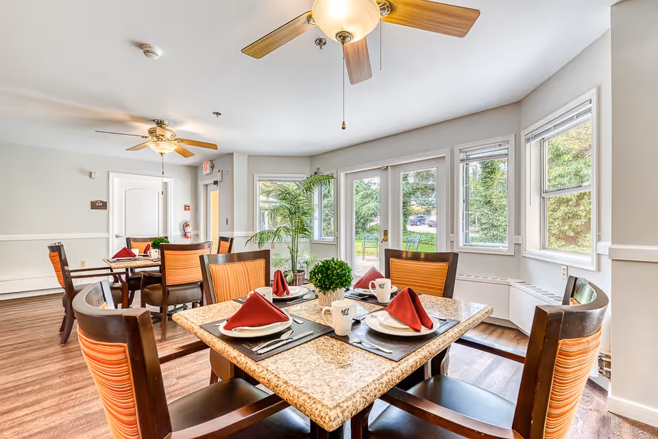 A bright dining room with multiple tables set for four people each, featuring granite tabletops, wooden chairs with orange-striped cushions, red folded napkins, white plates, cups, and silverware. The room has large windows and glass doors letting in natural light, ceiling fans, and a view of greenery outside.