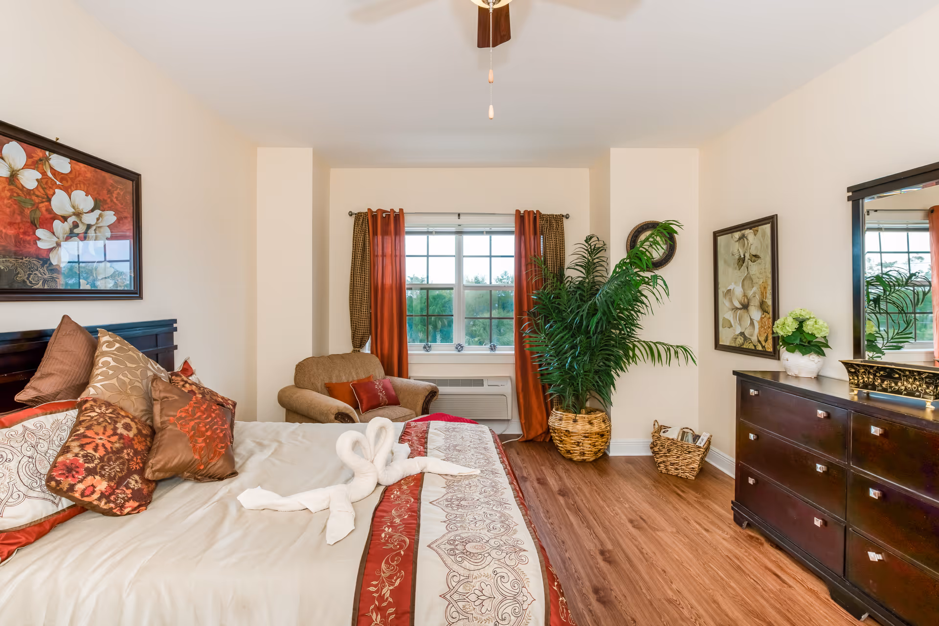 A well-decorated bedroom with a large bed adorned with multiple decorative pillows and a folded towel shaped like swans on the bedspread. There is a comfortable armchair near a window with red and gold curtains. A large green potted plant is placed next to a dark wooden dresser with a mirror above it. The room has wooden flooring and light-colored walls with floral artwork.