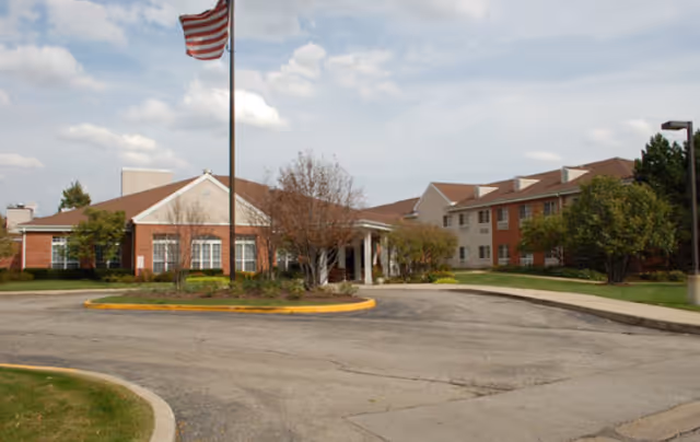 Exterior view of a senior living facility building with a circular driveway, an American flag on a flagpole, and landscaped greenery including trees and bushes under a partly cloudy sky.