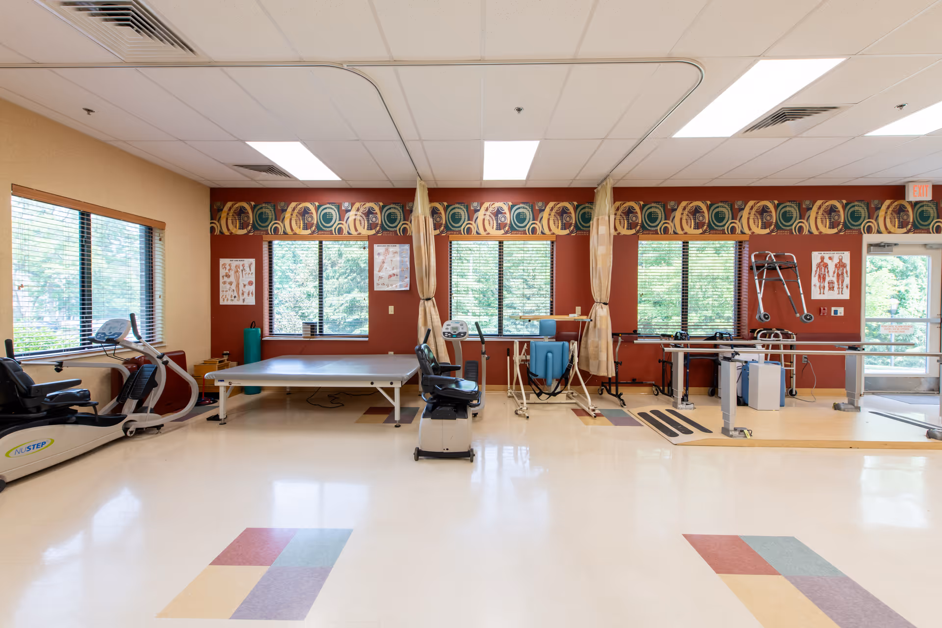 A spacious physical therapy room with exercise equipment including stationary bikes, parallel bars, a therapy table, and a walker. The room has large windows with blinds, colorful patterned valances, and anatomical charts on the walls. The floor has a light-colored surface with colored square patterns.
