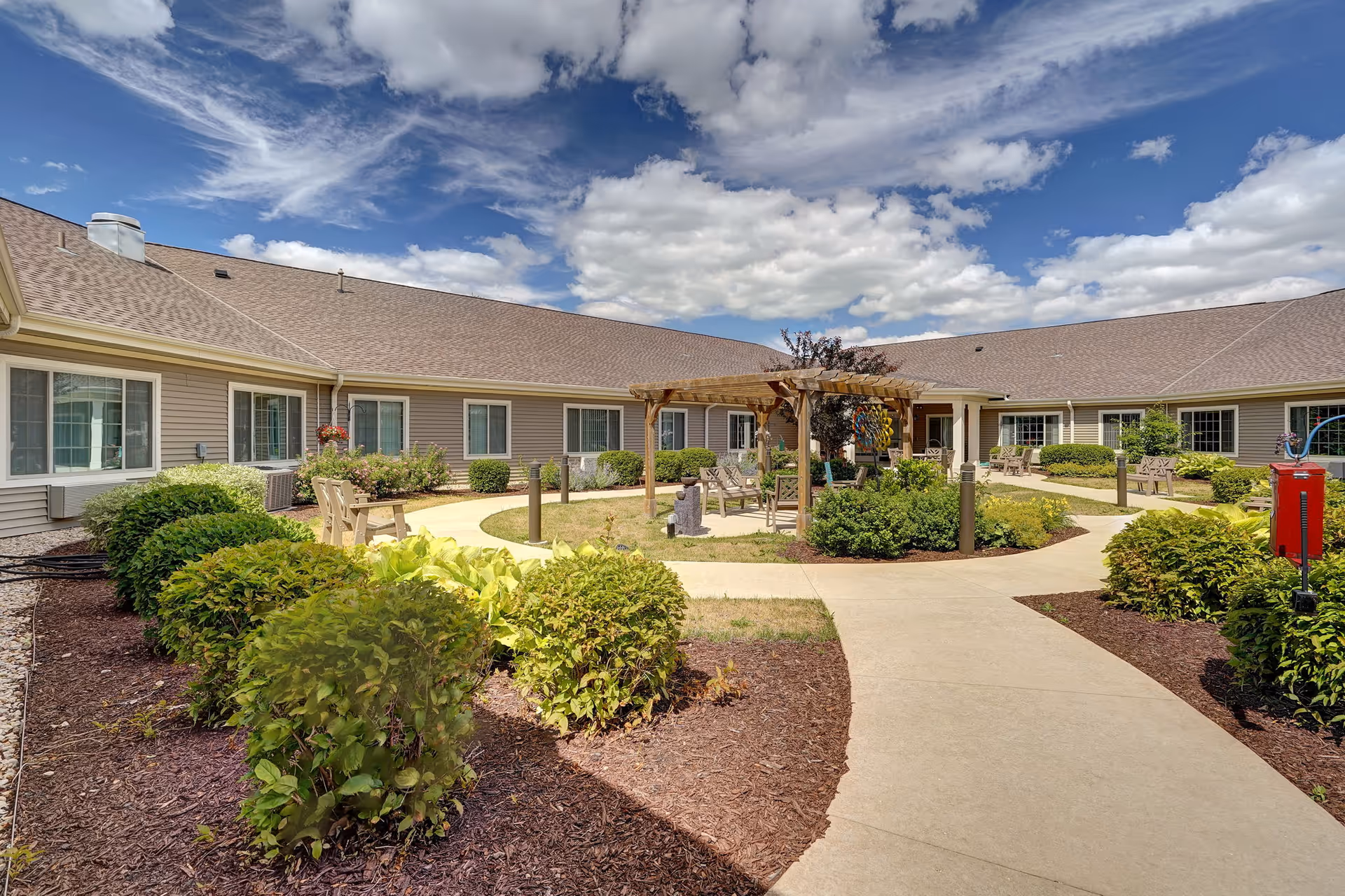 Outdoor courtyard area of a senior living facility with a paved walkway, green bushes, and a wooden pergola with benches underneath. The building surrounds the courtyard with multiple windows and a brown shingled roof under a partly cloudy blue sky.