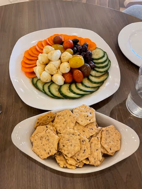 A table with a platter of sliced cucumbers, cherry tomatoes, mozzarella balls, olives, carrots, and a bowl of hexagonal crackers.