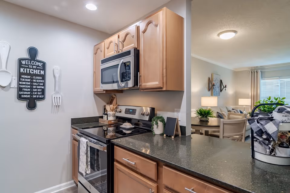 A modern kitchen with wooden cabinets, a black countertop, a stainless steel microwave and stove. On the wall, there is a decorative sign shaped like a cutting board with kitchen rules. The kitchen opens into a living area with a round table, chairs, lamps, and a window with blinds and curtains.
