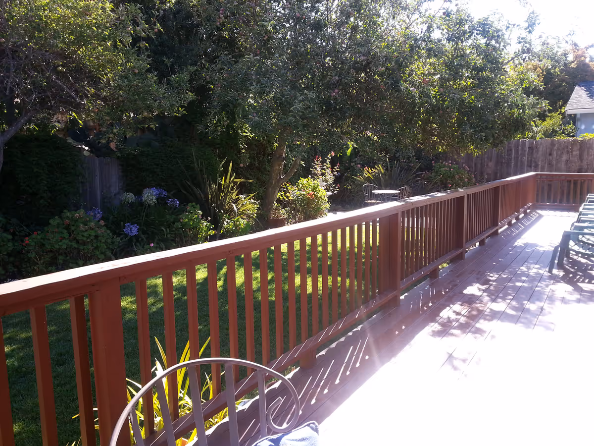 A sunlit outdoor deck area with wooden railings overlooking a green garden with trees, bushes, and flowering plants. There are metal chairs and a small table on the deck and in the garden area.