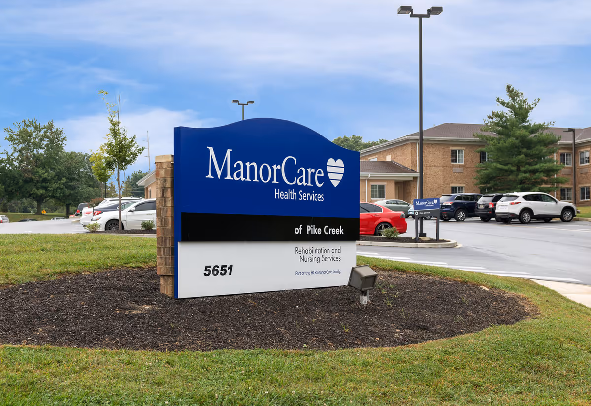 Outdoor view of the ManorCare Health Services sign for Pike Creek Rehabilitation and Nursing Services, located on a grassy area near a parking lot with several cars and a brick building in the background under a partly cloudy sky.