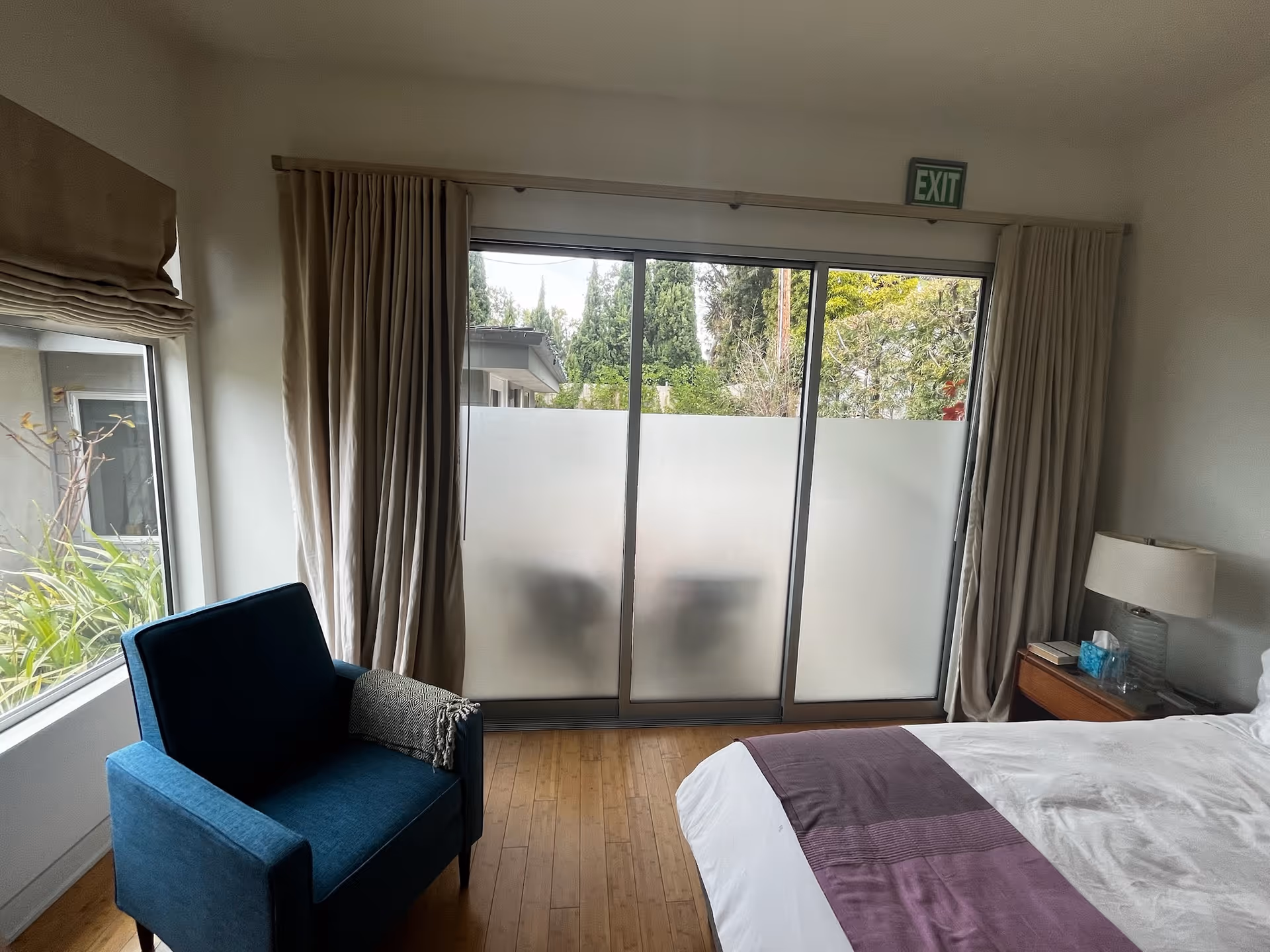 A bedroom with a bed partially visible on the right side, covered with white and purple bedding. Next to the bed is a wooden nightstand with a lamp, books, and a box of tissues. On the left side, there is a blue armchair with a folded blanket draped over one arm. The room has wooden flooring and large sliding glass doors with frosted lower panels, leading outside. Beige curtains frame the glass doors, and an exit sign is mounted above them. A window with a beige Roman shade is visible on the left wall, showing greenery outside.