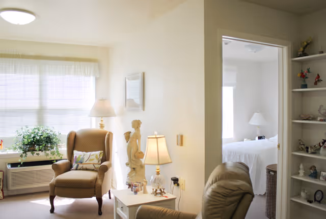 A cozy senior living room with a cushioned armchair, a side table with a lamp and a decorative statue, a large window with blinds letting in natural light, and a view into a bedroom with a white bedspread and a bedside lamp. There is also a built-in shelf with various decorative items.