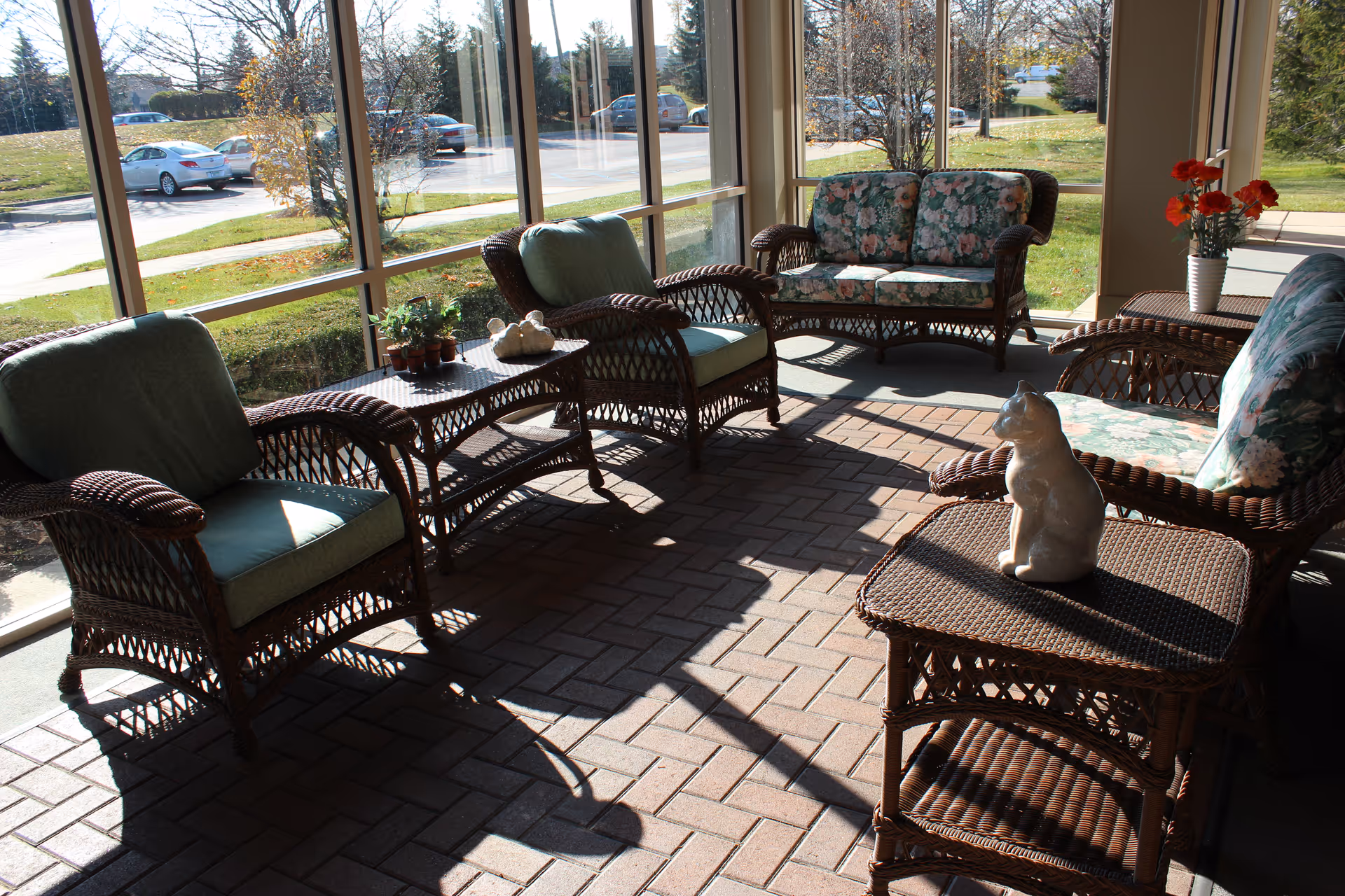 Sunlit enclosed patio/sunroom with wicker chairs and tables, floral cushions, potted plants and a ceramic cat, overlooking a lawn and parking lot.