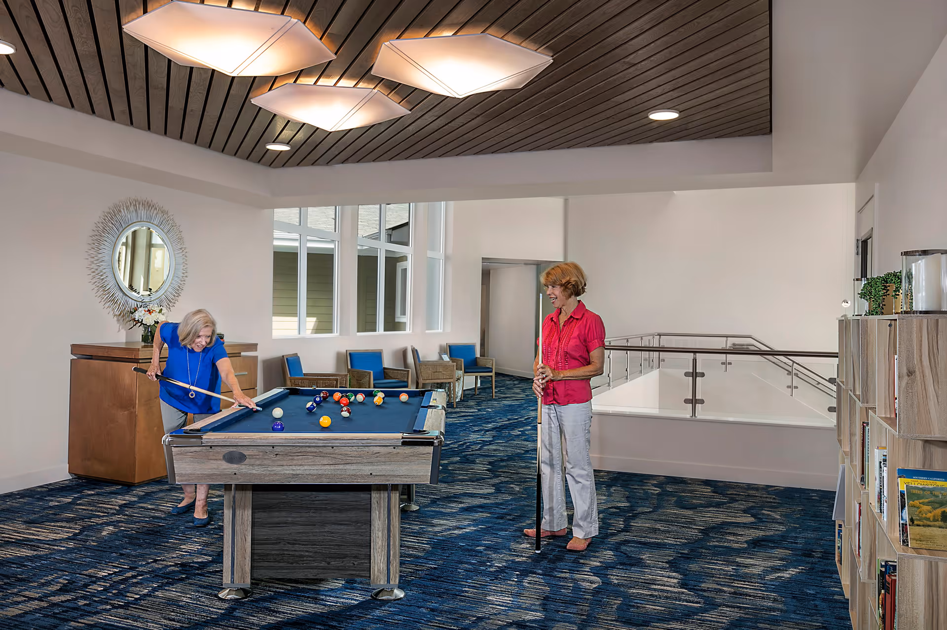 Two elderly women playing pool in a well-lit recreational room with blue carpet, modern ceiling lights, and large windows. One woman is taking a shot while the other watches. The room has chairs along the wall, a decorative mirror, and a bookshelf with books and plants.