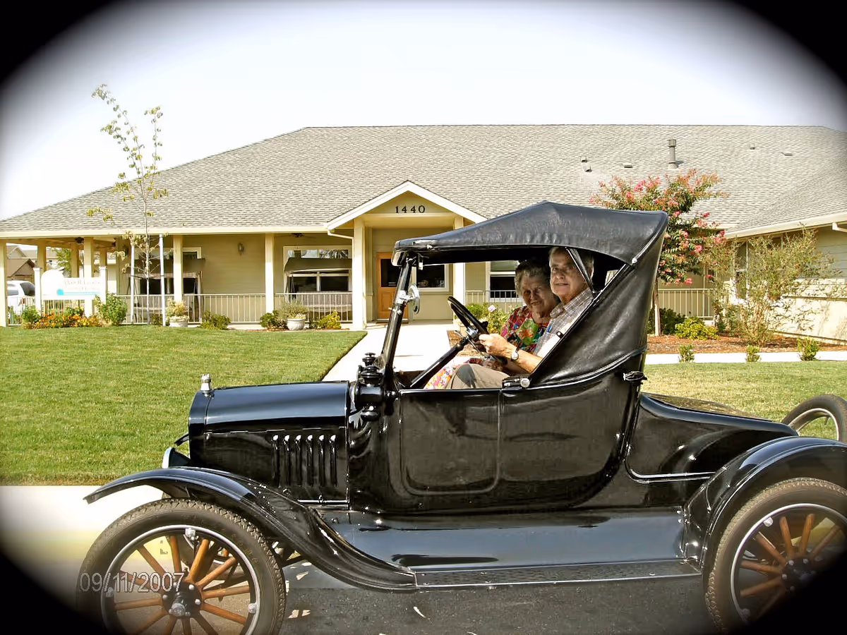An elderly couple sit in a vintage black car parked in front of the Westhaven Assisted Living building and its manicured lawn.