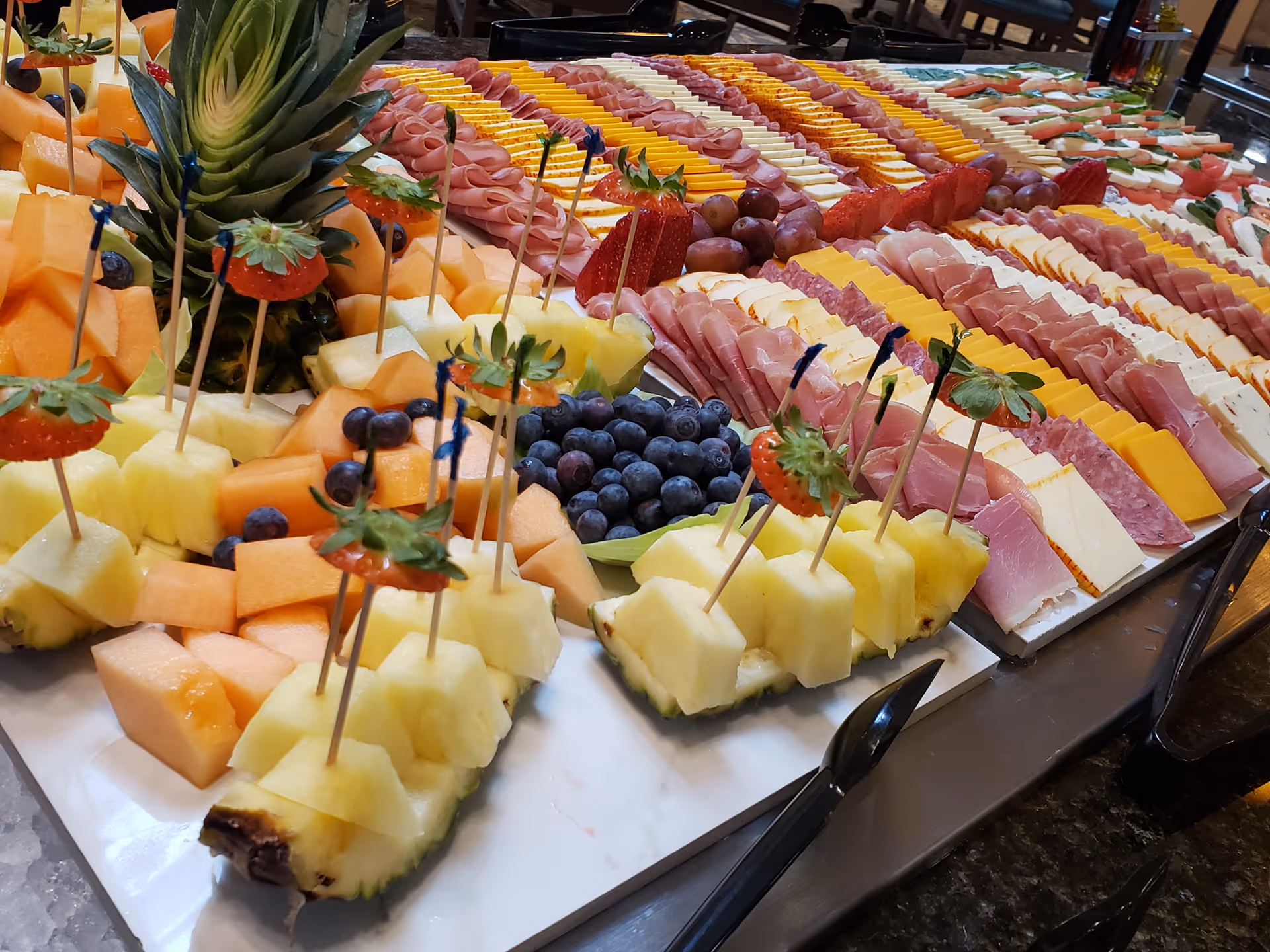 A buffet table with an assortment of sliced fruits including pineapple, cantaloupe, blueberries, and strawberries on skewers, alongside neatly arranged slices of various cheeses and deli meats.