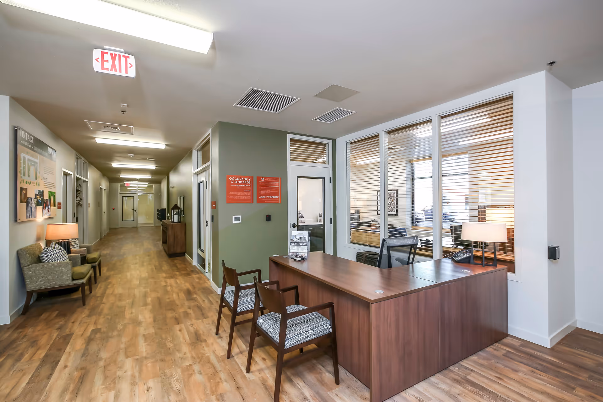 Interior view of a senior living facility hallway with wood flooring, a reception desk with two chairs, and a seating area with two armchairs and a lamp. The hallway extends into the distance with multiple doors and overhead lighting. There are large windows with blinds behind the reception desk.
