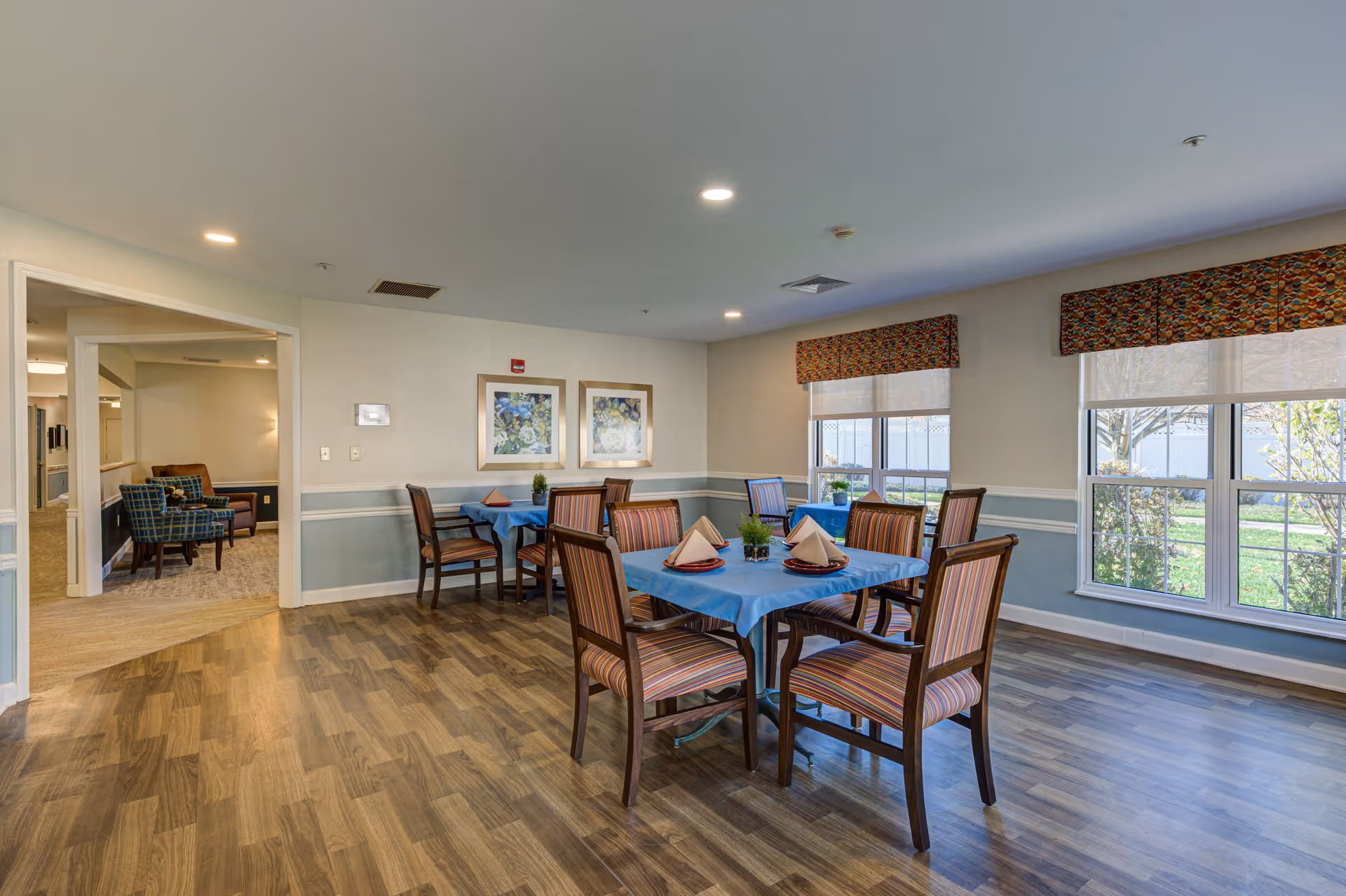 A dining area in a senior living facility with two tables covered in blue tablecloths, each set with folded napkins and small potted plants. The room has large windows with patterned valances letting in natural light, wood flooring, and framed floral artwork on the walls. Adjacent to the dining area is a lounge space with upholstered chairs.