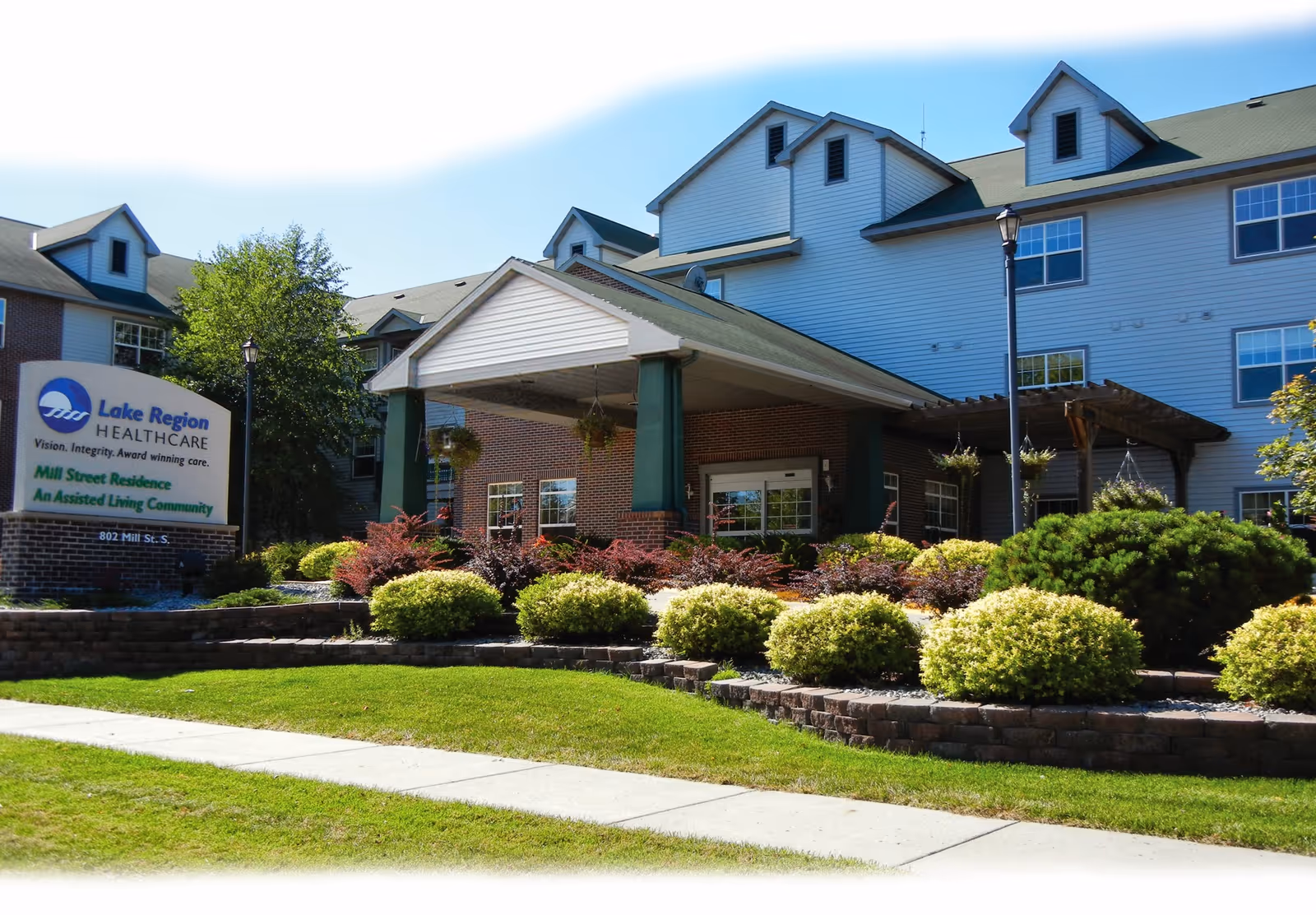 Exterior view of Mill Street Residence, an assisted living community, showing a multi-story building with a covered entrance, well-maintained landscaping with bushes and trees, and a sign for Lake Region Healthcare.