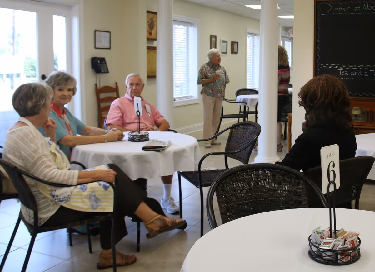 A group of elderly people sitting and chatting around tables with white tablecloths in a bright dining area. One woman stands near the back holding a cup, and another person is partially visible in the background. The room has large windows, white walls, and a blackboard with writing on it.