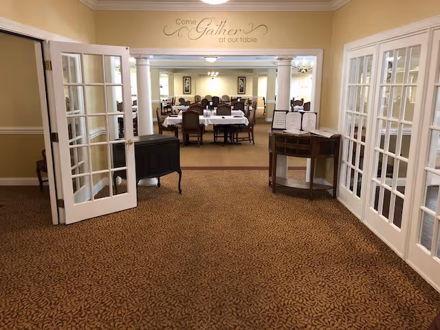View through open double glass doors into a dining room with multiple tables covered with white tablecloths and surrounded by chairs. The walls are painted beige, and there is a decorative sign above the doorway that reads 'Come Gather at our table'. The carpet has a patterned design, and there are small tables with menus or papers near the entrance.