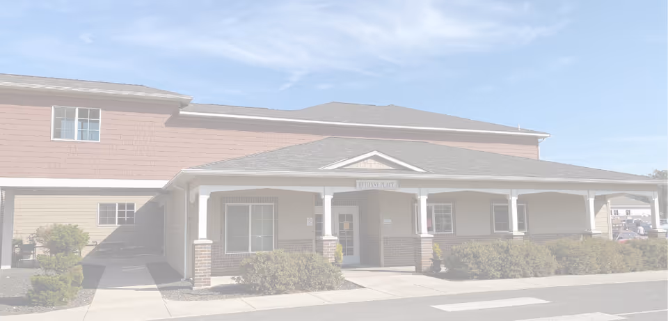 Front exterior of a two-story senior living building with a covered entrance, white columns, and low shrubs.