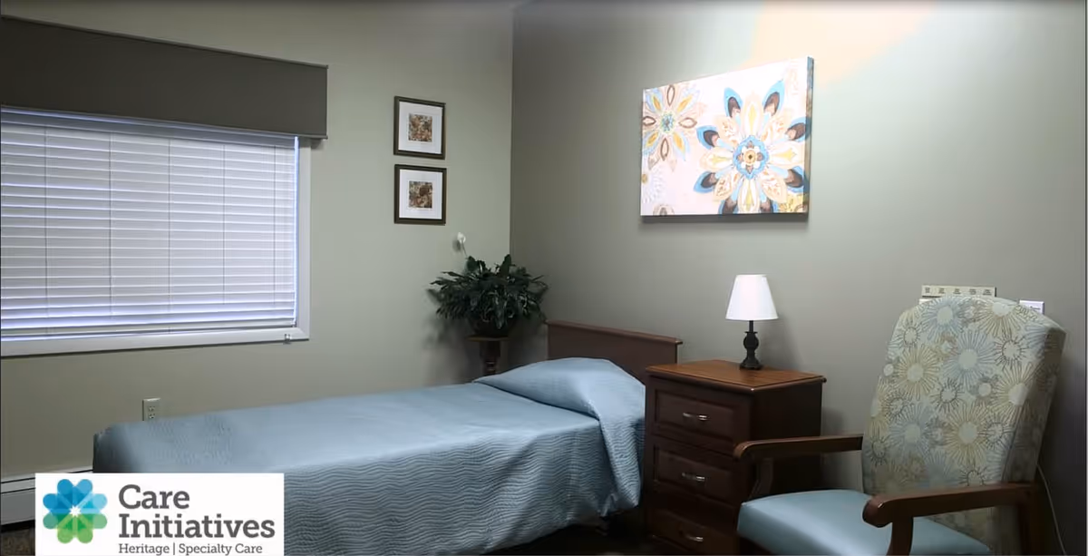 A small, neatly arranged bedroom with a single bed covered in a light blue bedspread. Next to the bed is a wooden nightstand with a small lamp. A cushioned armchair with floral upholstery is positioned beside the nightstand. The walls are painted a light grayish-green and decorated with two small framed pictures and a larger floral artwork. A window with closed blinds is on the left side of the room.