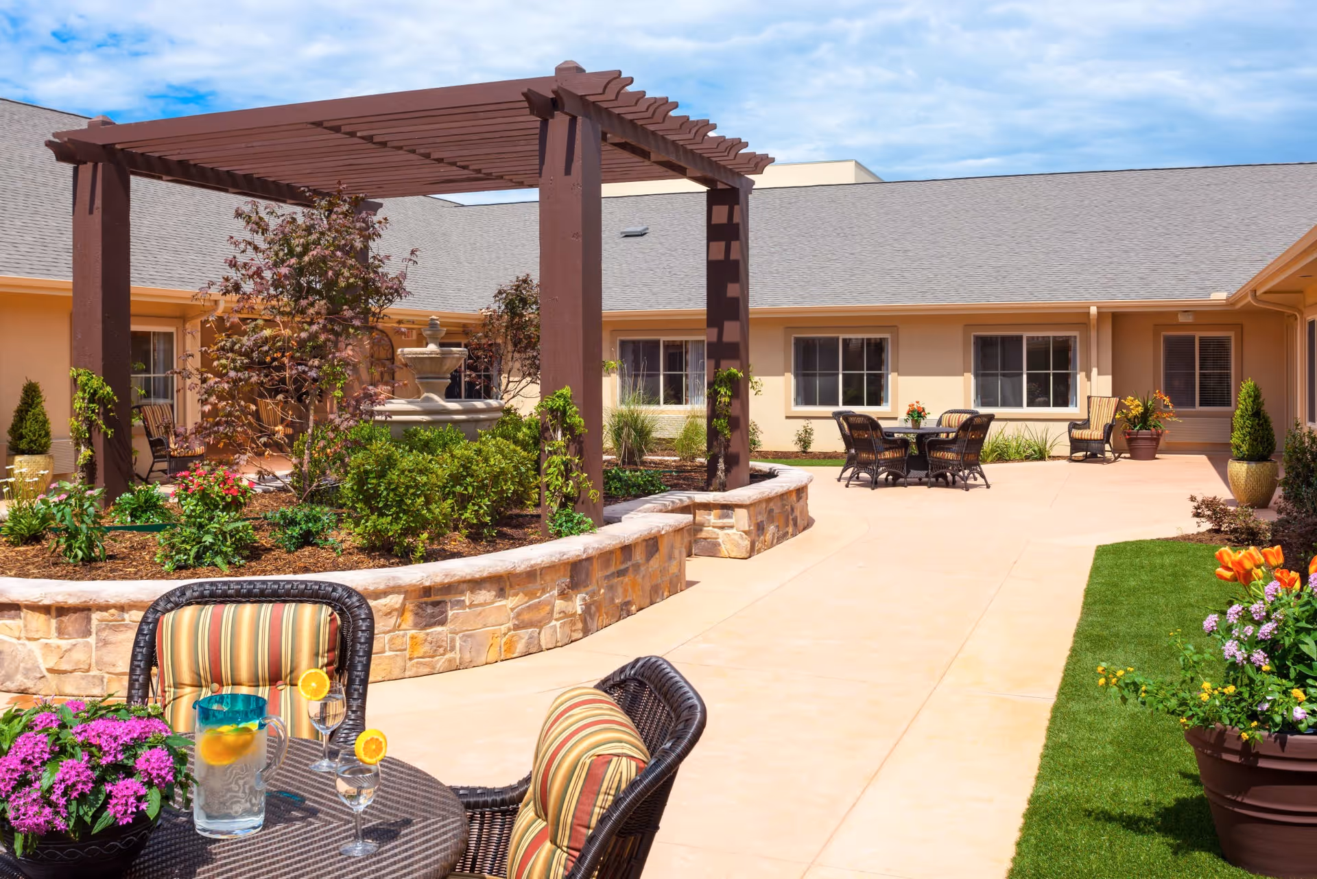 Outdoor courtyard area at Riverside Oxford Memory Care featuring a stone planter with a wooden pergola, various plants and flowers, patio tables and chairs with striped cushions, and a water fountain in the background under a partly cloudy sky.