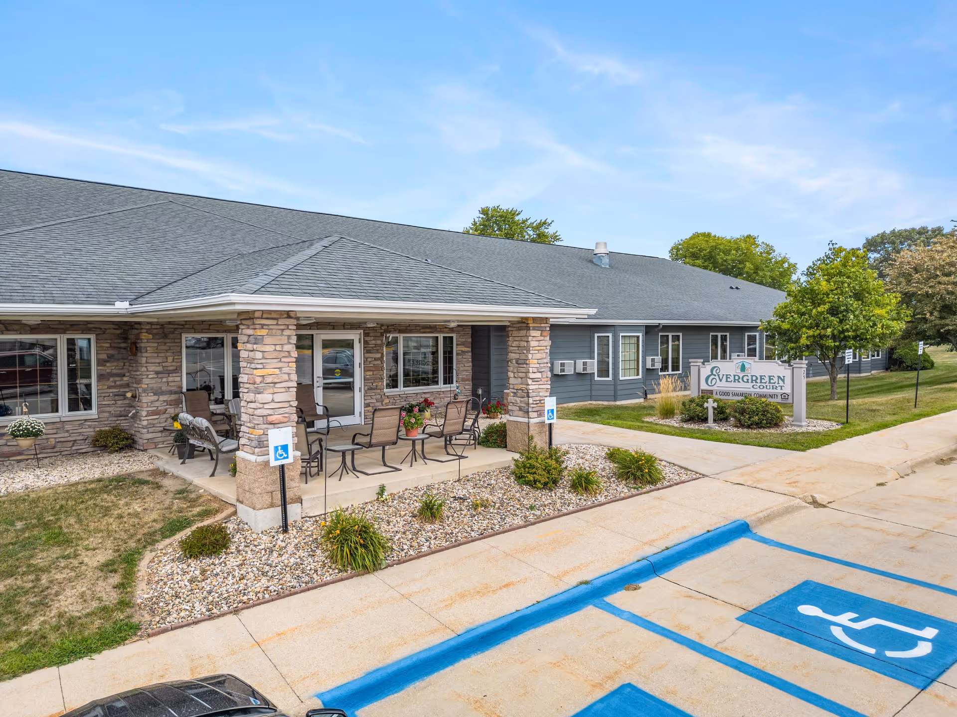Exterior view of a single-story senior living facility building with a covered patio area furnished with chairs and tables. The building has stone pillars and blue-gray siding. There are landscaped areas with small bushes and plants, a sidewalk, and marked handicap parking spaces in the foreground. A sign near the building reads 'Evergreen Court, A Good Samaritan Community.'