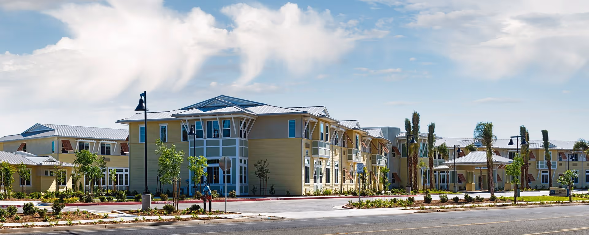 Exterior view of a large, modern senior living facility building with multiple windows, palm trees, and landscaped greenery under a partly cloudy sky.
