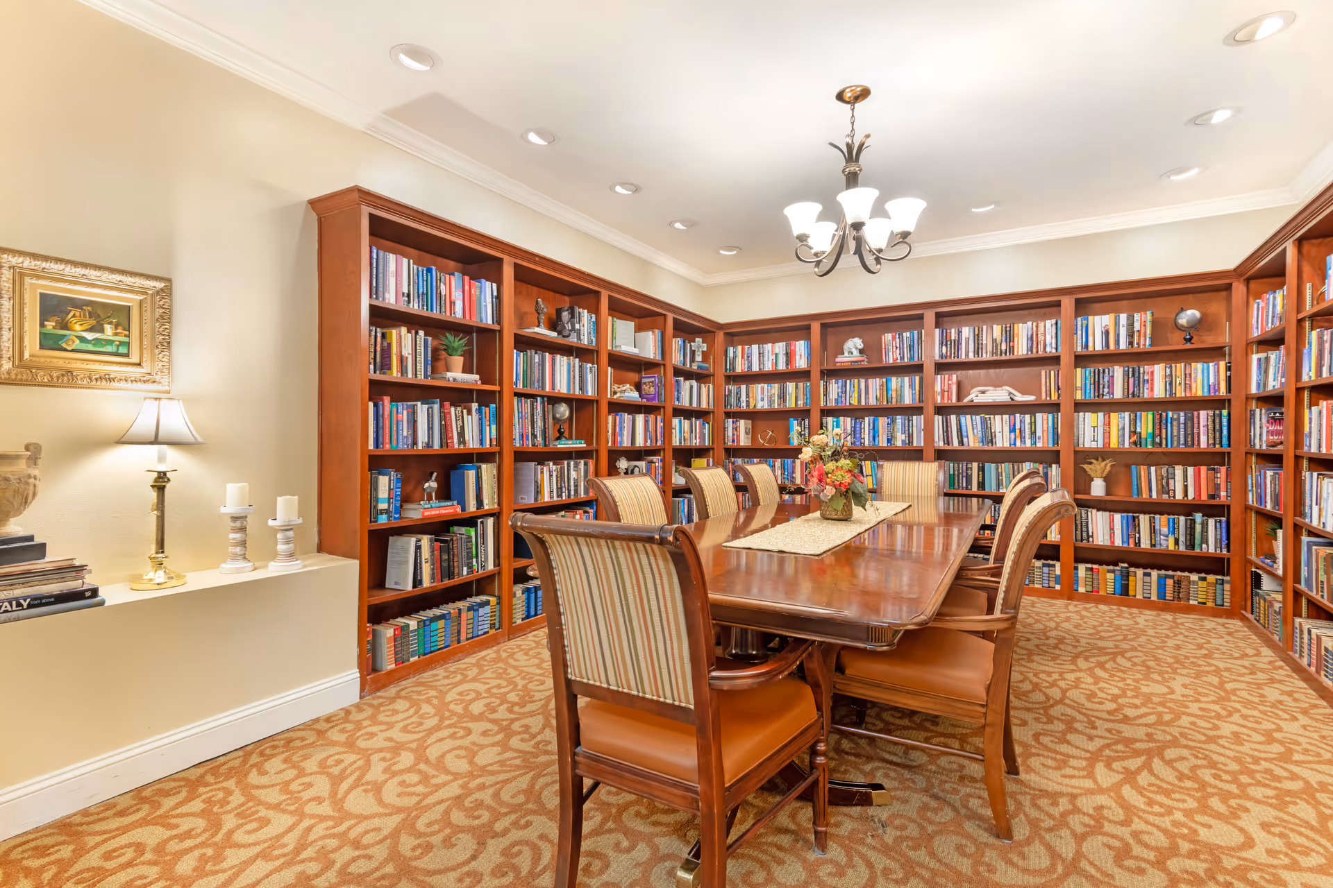 A well-lit library room with wooden bookshelves filled with books lining the walls. In the center, there is a large wooden table surrounded by eight upholstered chairs. A chandelier hangs from the ceiling, and a small table lamp and decorative items are placed on a shelf on the left wall. The carpet has a patterned design in warm tones.
