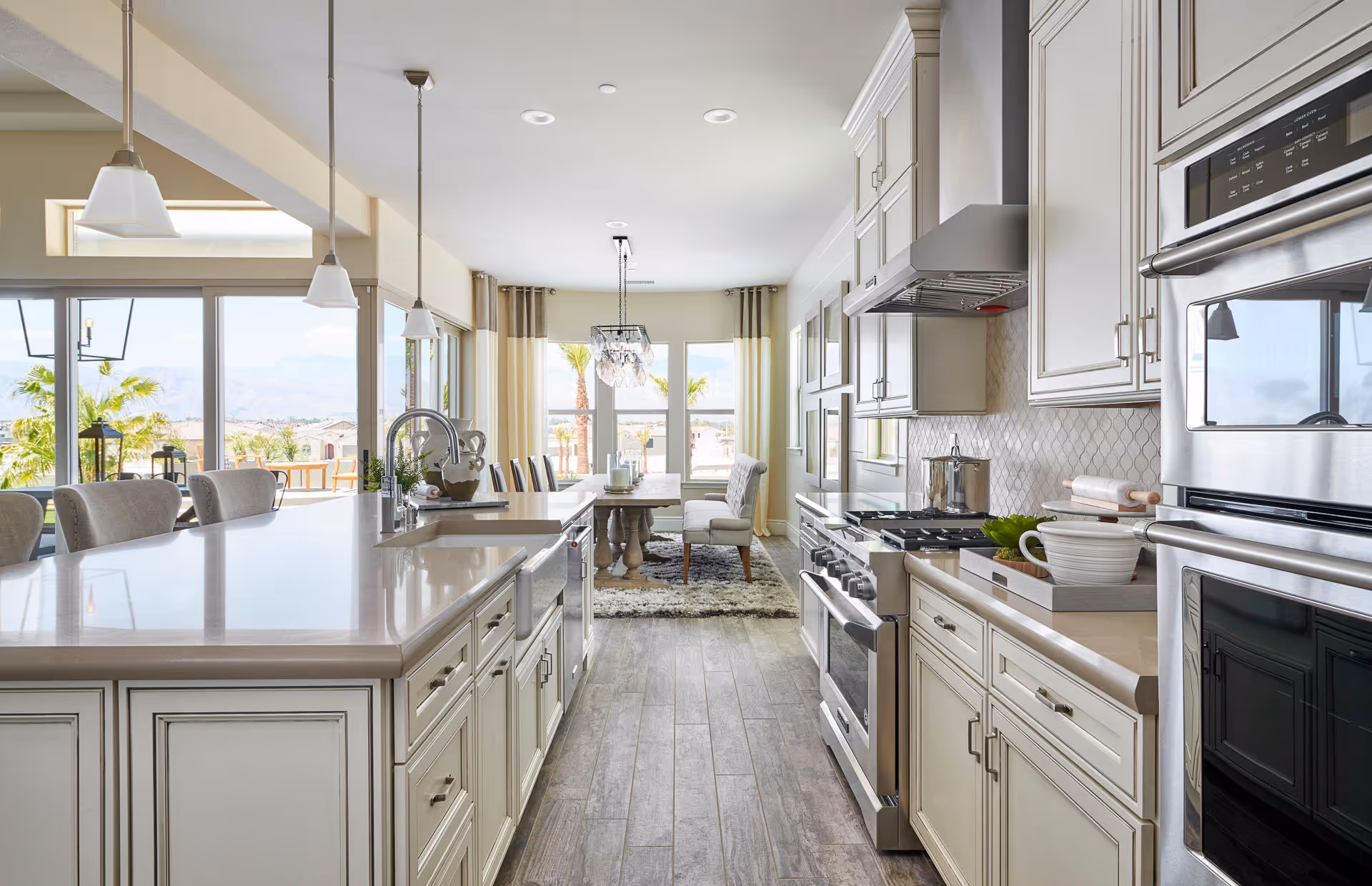 Bright and spacious kitchen with white cabinetry, stainless steel appliances including a stove and double oven, a large island with a sink and seating, pendant lights hanging above, and a dining area with a table and chairs near large windows showing an outdoor view with palm trees and mountains.