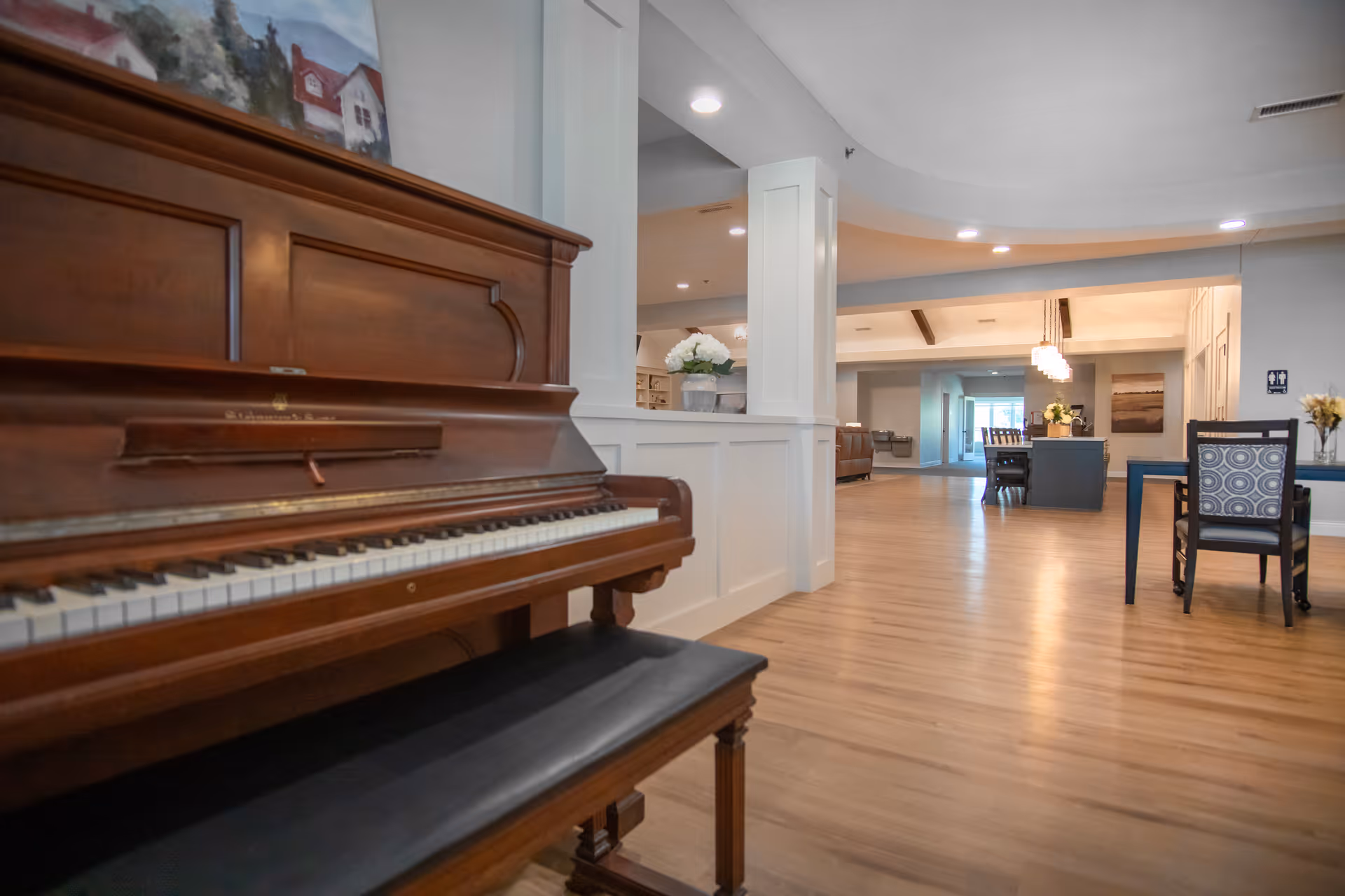 Interior view of a senior living facility with a wooden upright piano and bench in the foreground on the left. The room features light wood flooring, white walls with wainscoting, and recessed ceiling lights. In the background, there are tables and chairs, a flower arrangement on a table, and a hallway leading to other areas of the facility.