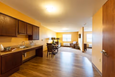 Interior view of a senior living facility room with a kitchenette on the left featuring a sink and cabinets, a dining table with chairs in the middle, and a living area with armchairs and a TV near the windows in the background. The walls are painted yellow and the floor is wood and carpeted in the living area.