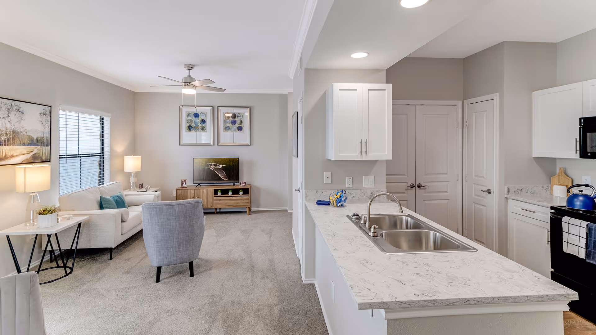 A bright and modern open-concept living room and kitchen area. The living room features a beige sofa with blue cushions, a gray armchair, a side table with a lamp, and a TV on a wooden stand against a light gray wall with framed artwork. The kitchen has white cabinets, a marble countertop with a double sink, and black appliances including a stove with a blue kettle. The space is carpeted and well-lit with natural light from a window and ceiling lights.