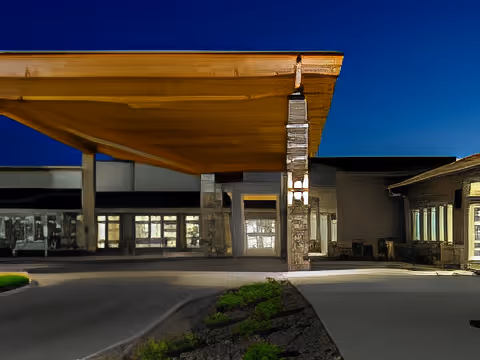 Exterior view of a building at night with a large wooden canopy extending over the driveway entrance, illuminated by lights on stone pillars. The building has multiple windows and a modern architectural style.