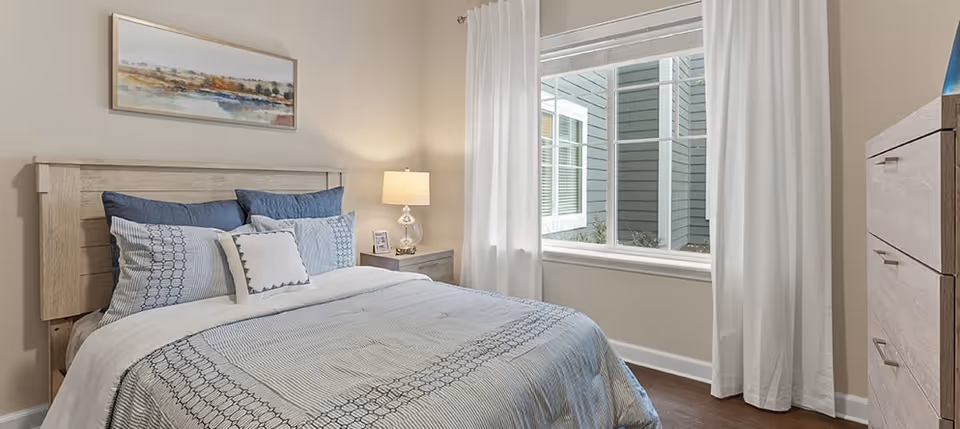 Sunlit bedroom with a made bed, nightstand and lamp, white curtains over a window, and a dresser.